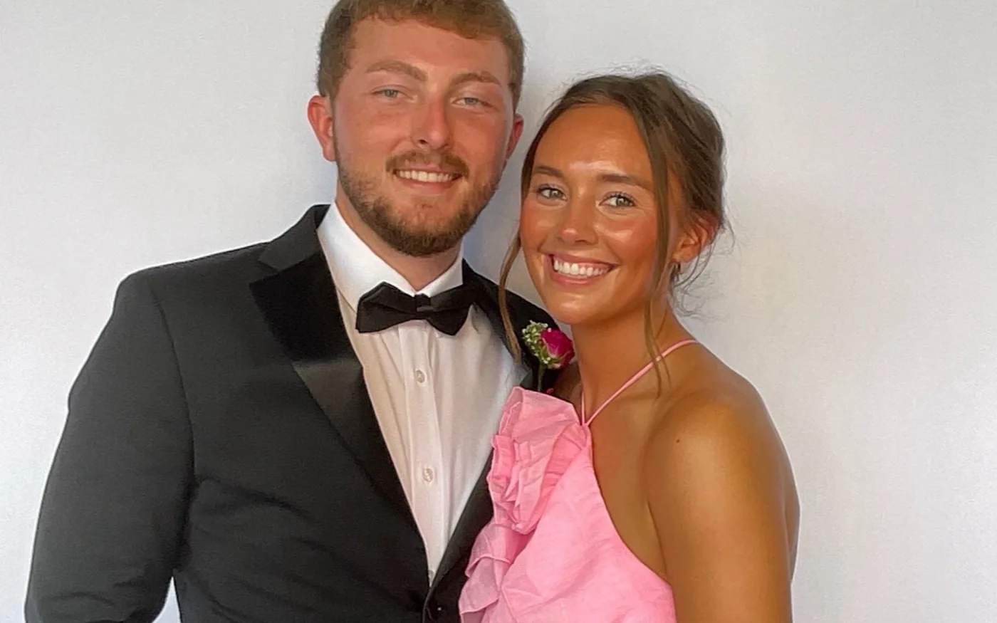 A couple in a tuxedo and a pink dress posing together in front of a white backdrop at a wedding