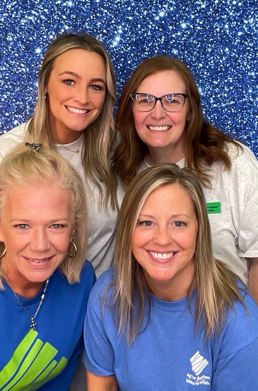 Four women in matching event t-shirts smiling together in front of a blue glitter backdrop