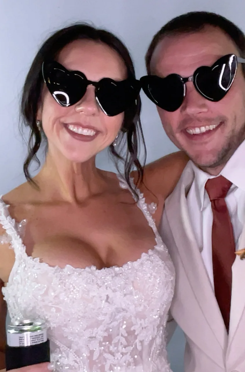 A bride and groom in heart shaped sunglasses smiling together in the photo booth