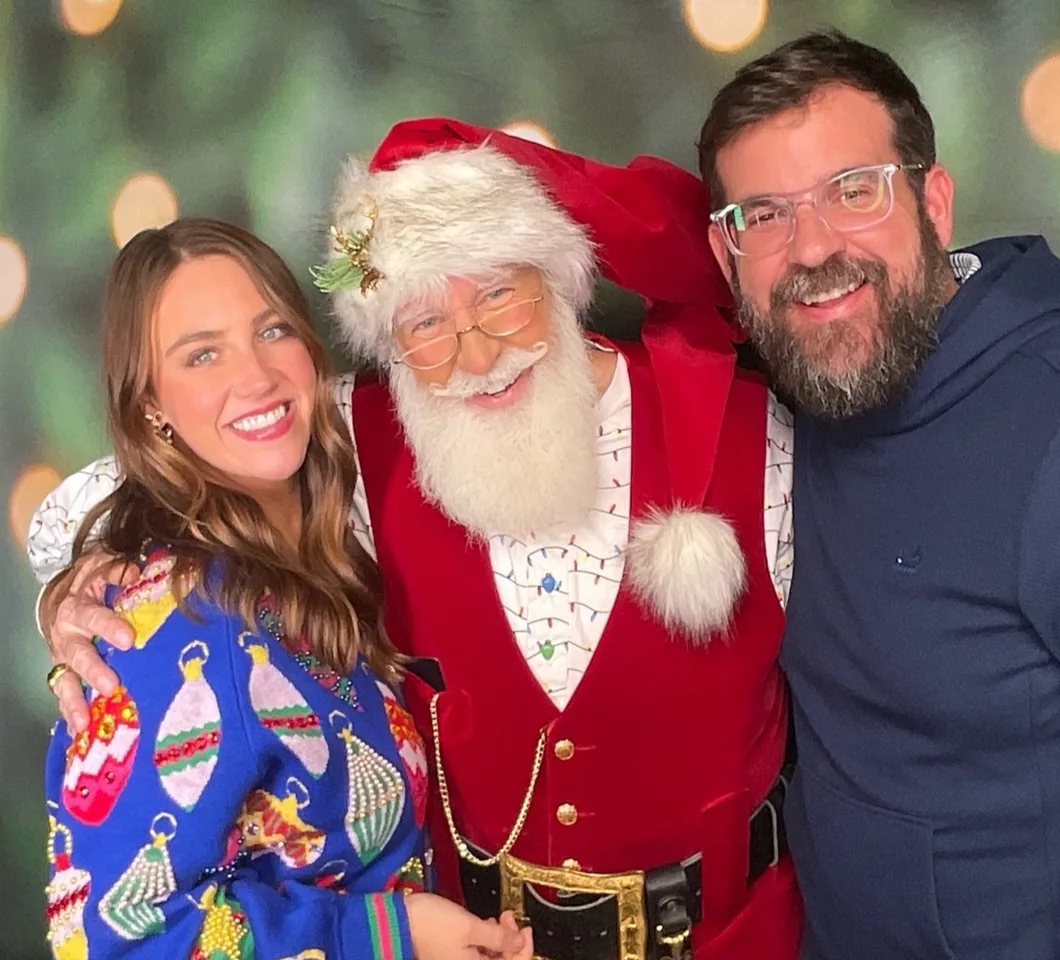 Two guests either side of Santa in the booth, in front of a Christmas tree backdrop